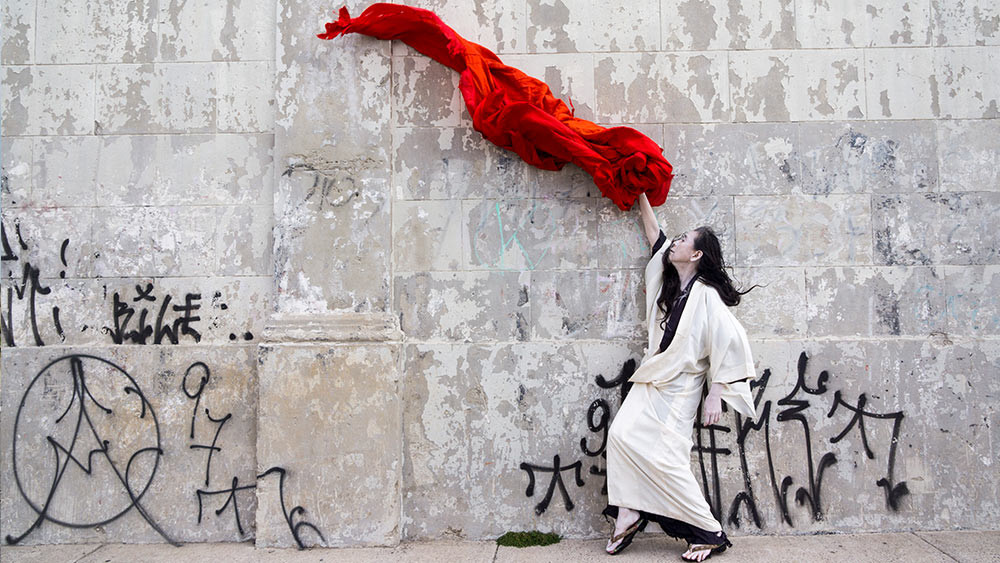 Eiko Otake makes a gesture, while standing infront of a white wall with black grafitti dressed in a white and black kimono, which sends a piece of red fabric flying overhead. Photo by William Johnston