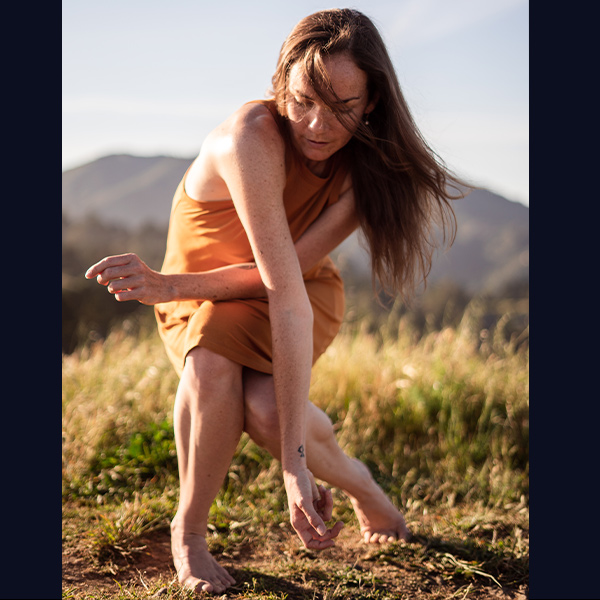 Casey Thorne dances outside, feet on bare ground, surrounded by tall grasses and a mountain on the horizon. Dressed in an orange dress, their left knee locks in behind a deeply bent right leg, with their right arm draped long over the left, reaching for the ground.