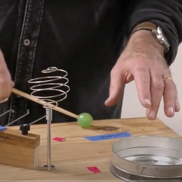 An array of objects sit on a wooden board: a upside down spiral whisk, a portion of a coffee caen. A green rubber mallet is striking the board near some blue and pink tape. a left hand floats sensitively over the board. Photo still from video.