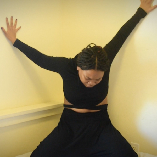 Dancer Vu Mbambo stands in a corner, dressed in black with her hair pulled back in braids, with both arms stretched up between the walls and her head down
