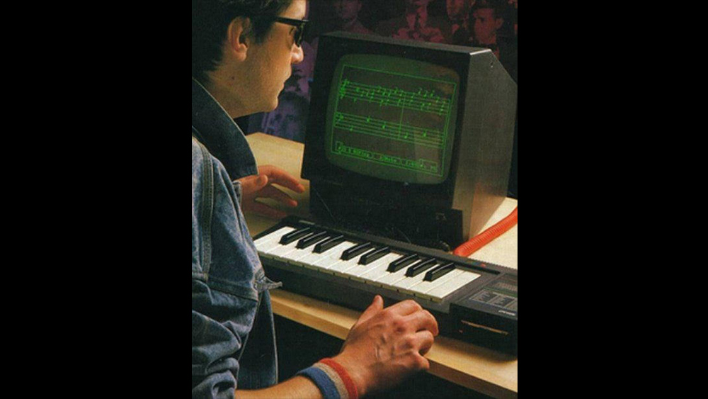 A retro photo (80's-ish) of a person in a jean jacket, collar up with red white and blue wrist bands sitting in front of a small two octave keyboard with an old monitor displaying musical staves in green led light.