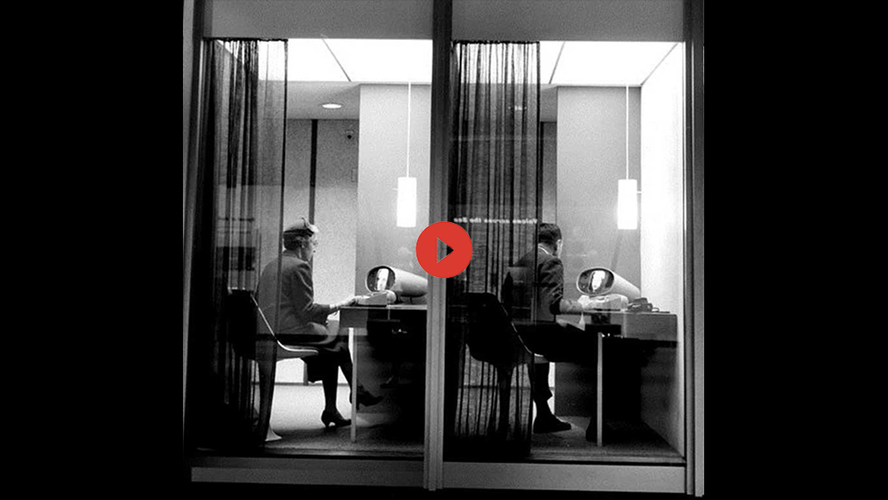 a black and white image of two people, a man and a woman, sitting indoors looking into an early video call device mounted on a desk. The camera is observing the people through the window, their backs are turned to the viewer. 