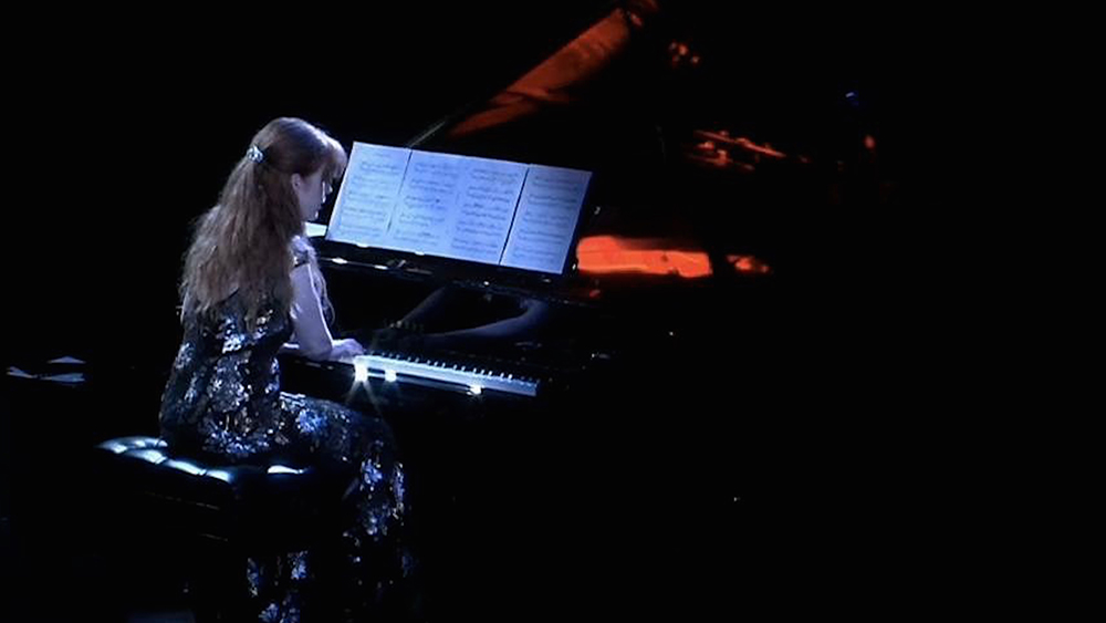 Sarah Cahill sits at a piano, dressed in a metallic embroidered dress. Phot courtesy of the Artist.