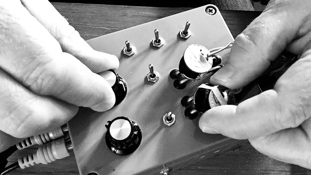 A close-up, black and white photo of fingers manipulating knobs on a small controller with four knobs and five switches.