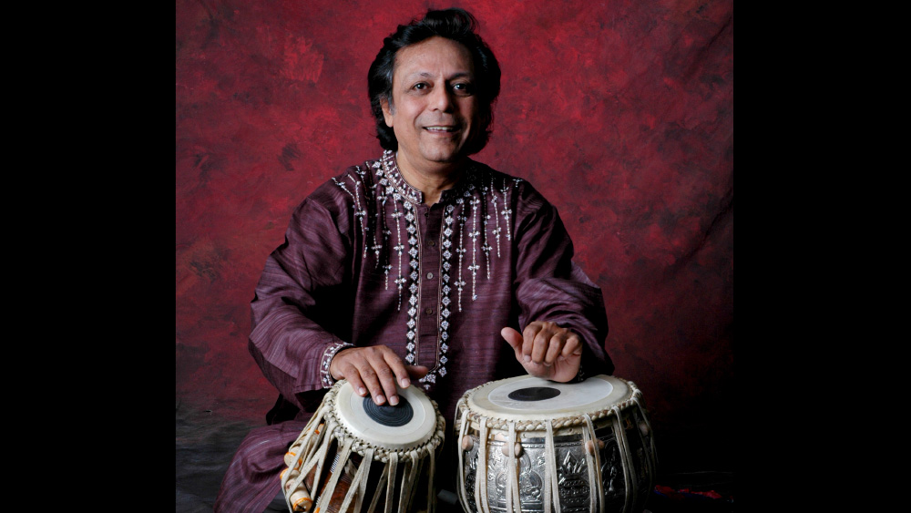 Swapan Chaudhuri is seated behind tabla drums, both hands paying the heads. They are dressed in a burgundy garment with white and black details on the collar and front. Swapan is facing that camera directly, looking to the viewer.