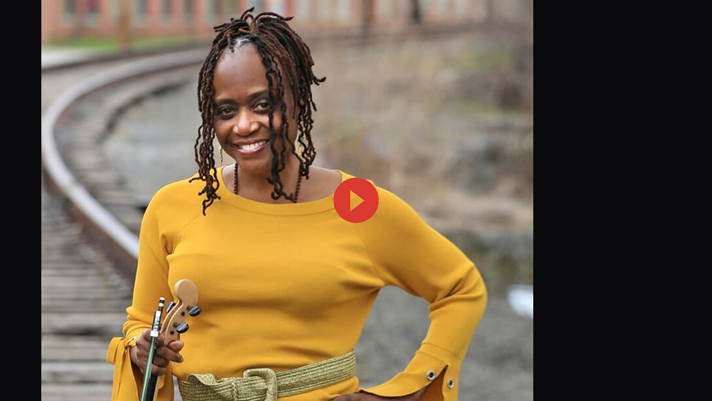 Gwen Laster stands in a railroad yard, wearing a yellow sweater and holding their viola. They are smiling and looking at the camera.