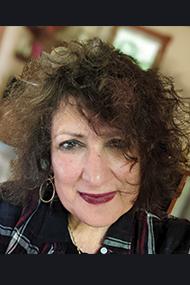 Author Elmaz Abinder looks into the camera, dark curly hair framing their face.