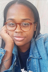 Author Janae Newslon looks into the camera, their head resting on their right hand, dressed in a jean jacket and white t shirt.