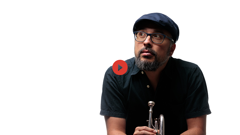 Taylor Ho Bynum sits infront of a white background, looking up to the right on an angle through rimmed glasses, out from under the brim of a blue driving cap. Their instrument is held in their hands. Photo by Ben DeFlorio.