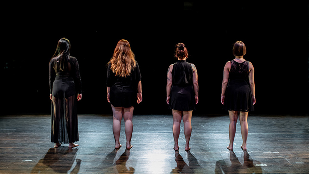 A color photograph with four bodies standing on a dark stage under theatrical light, back to the camera, looking out into a black void. Photo by Stephen Texeira.