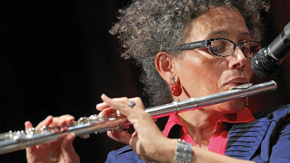 Nicole Mitchell plays flute, dressed in a blue shirt with a red earring. Photo Courtesy of the Artist.