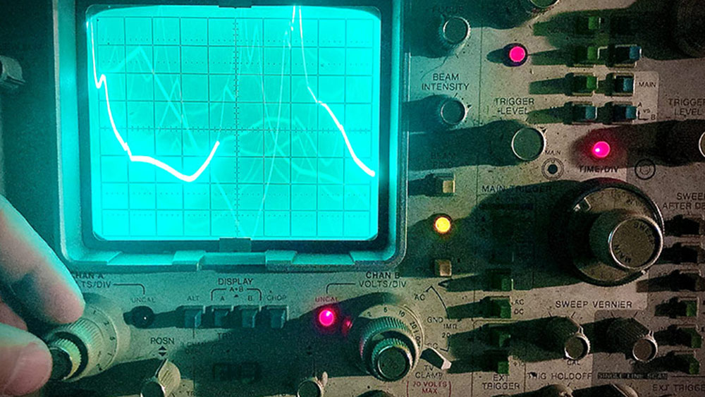 A photo of the face of an oscilloscope, featuring many knobs and buttons of various sizes, three red legs and a single yellow one, with a bleed green screen in the top left quarter being traced by a bright white line with peaks and valleys. Fingers and a thumb eating into frame from the left and touch a large knob. Photo by Jefferson Doyle.
