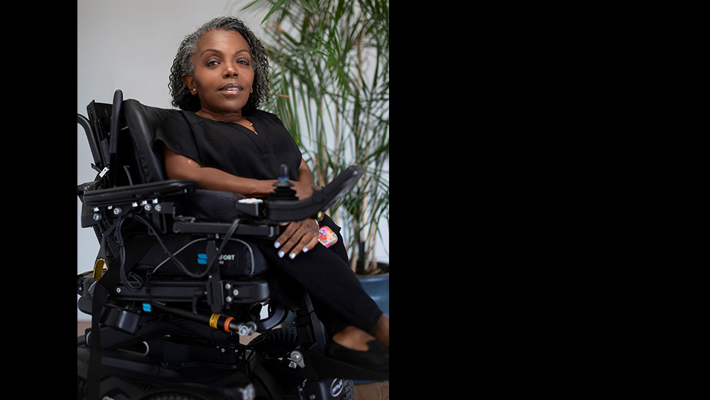 A color photgraph of Yomi Wrong, dressed in a black shirt, pants and shoes, seated in their wheelchair, looking into the camera wearing a gentle smile. Photo courtesy of Yomi Wrong.