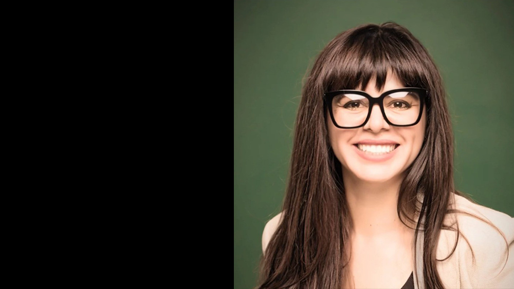 The image features a woman with long dark hair and full bangs, smiling brightly at the camera. She is wearing large, round black-rimmed glasses that frame her face, and her expression is cheerful and welcoming. The background is a muted green, providing a clean, solid contrast to her light skin tone and dark hair. Her outfit includes a beige blazer over a dark top, giving her a professional yet approachable appearance. The lighting is soft, highlighting her features and creating a warm, friendly atmosphere.