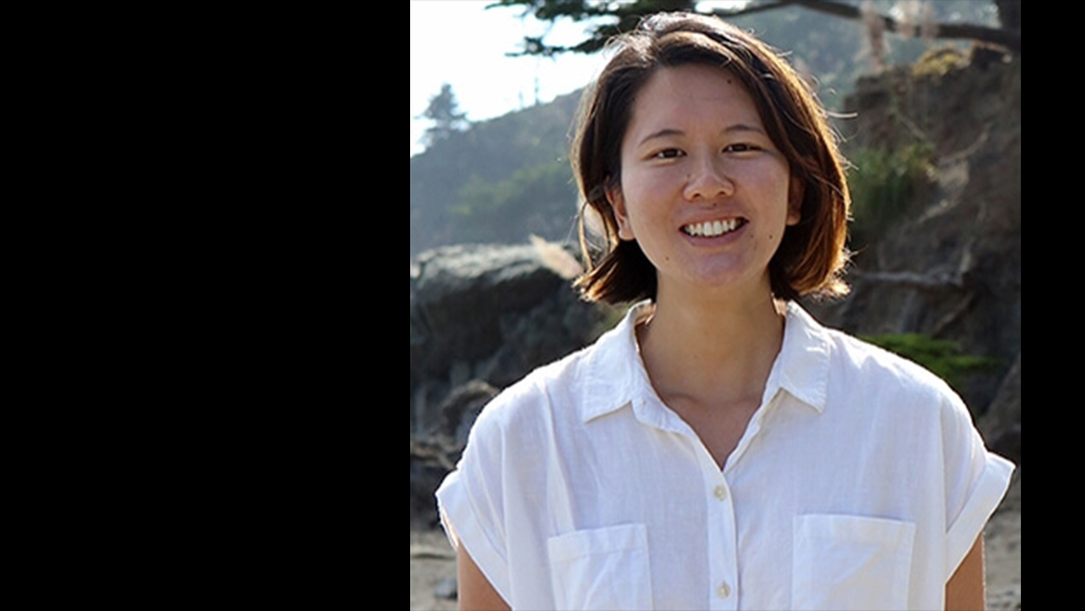 A color photograph of Heidi Lim, looking into the camera, their smiling face framed by chin length drak hair, dressed in a white shirt. Photo courtesy of Amelia Lim.