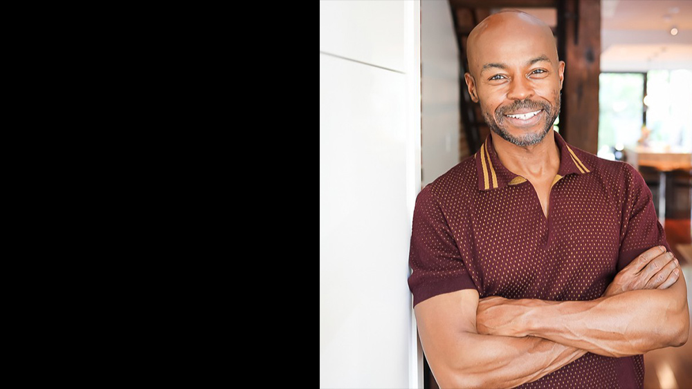 The color photgraph features a smiling man standing confidently with his arms crossed. He is wearing a short-sleeved, maroon-colored polo shirt with a subtle pattern and mustard accents on the collar. The background shows a warmly lit modern interior with blurred elements, such as a staircase and windows, suggesting a cozy and professional environment. The man appears approachable and self-assured.