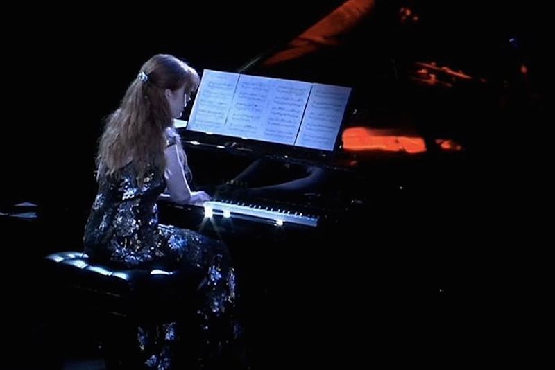 Sarah Cahill, dressed in a sparkling reflective gown, is seated at the piano with her back to the viewer. The sheets of music an the white keys of the piano are illuminated in a white light, while a red light shines into the body of the open grand piano. Photo courtesy of the Artist.