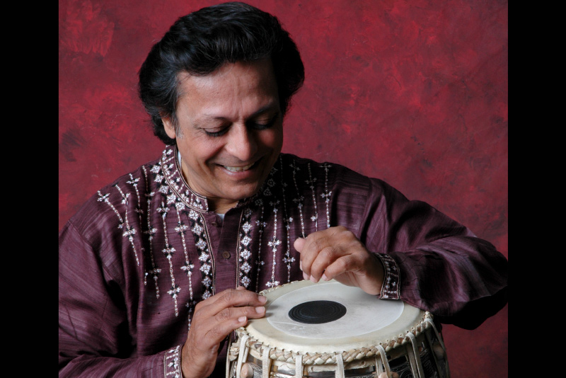 Swapan Chaudhuri, dressed in a burgundy garment, smiles while looking downward at their hands as they play a tabla drum.