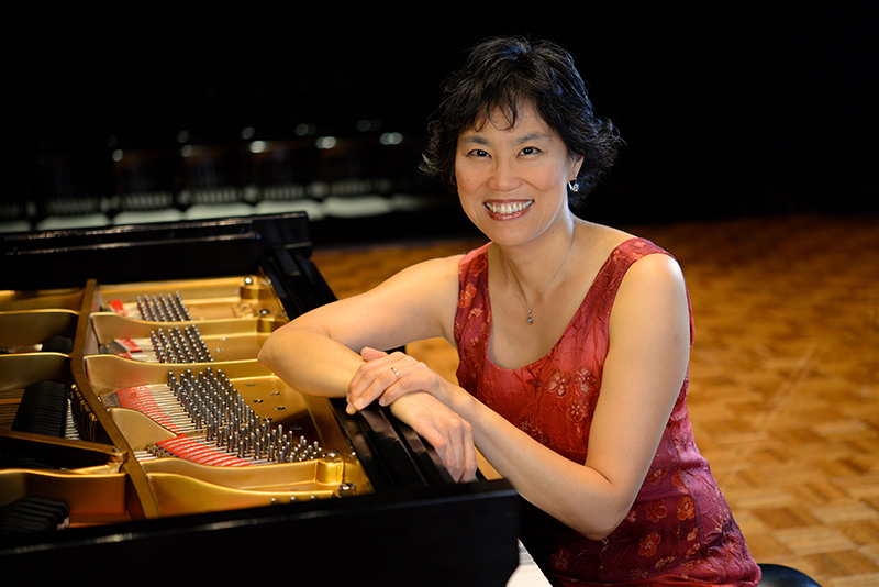 Genevieve Lee is seated, smiling into the camera, at the keyboard of a concert grand piano in a red silk dress with their hands folded atop the instrument. Photo courtesy of the Artist.