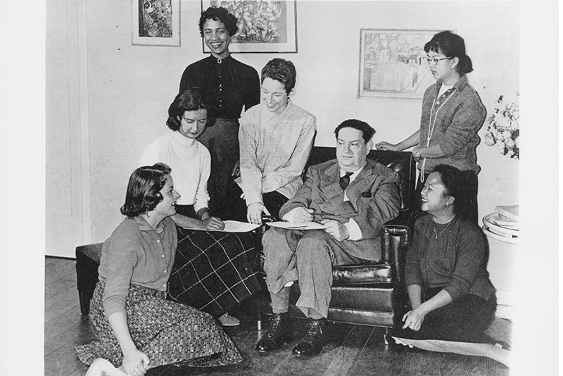 A black and white photograph of composer and teach Darius Milhaud, dressed in a grey suit and tie, with several women seated and standing around him, seated in an upholstered chair. Photo by Don Jones.  