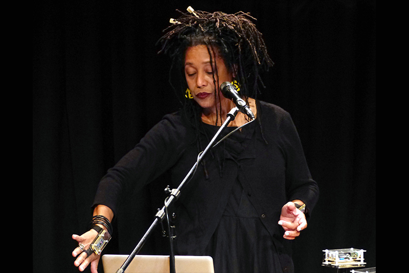 Pamela Z looks down to their extended right hand, which touches a table with electronic equipment and computers. A microphone stands before them. Their left hand is held aloft in a gentle gesture.