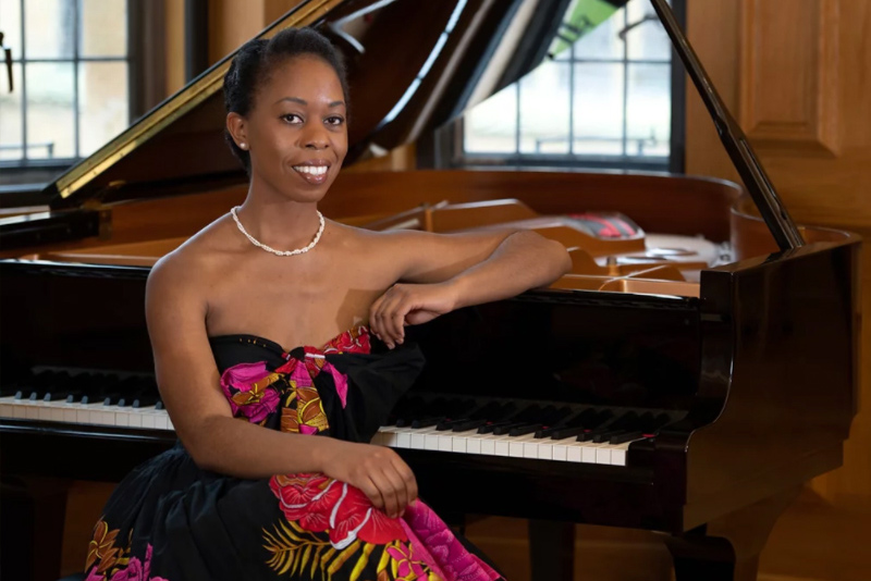 Samantha Ege is seated at the keyboard of an open grand piano in a wood paneled room. Dressed in a black dress with pink and gold floral print, she smiles broadly, her left elbow resting on the top of the glossy black piano. Photo by Jason Dodd.