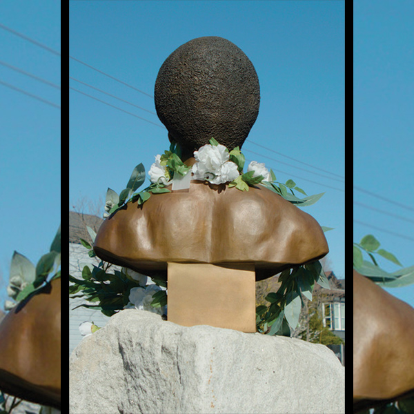 A color photograph of the Newton monument in Oakland CA featuring a sculted bust featuring a broad, muscled back and shoulders and the back of a head wearing short cropped hair, the scultpure is adorned with white flowers. Image courtesy of the filmmaker.