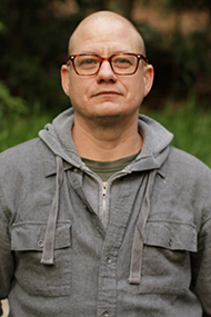 Cary Cronenwett looks directly into the camera through tortoise shell rimmed glasses, dressed in a grey hooded sweatshirt and grey jacket, a gentle smile on their face. Photo courtesy of the artist.