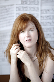 Sarah Cahill looks into he camera, both hands near her head tighter holding a lock fo her red hair, seated before large sheets of musical score. Photo by Christine Alicino.