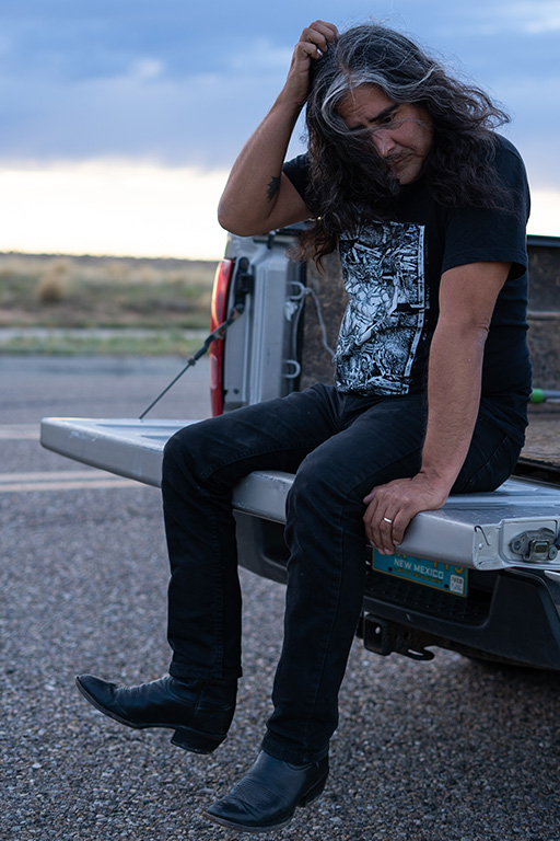 Artist Raven Chacon, dressed in a black t shirt and jeans, is seated in the back of a pick-up truck on the bed gate. They are leaning forward and looking downward, their long hair hanging as a hand reaches toward their head. The broad sky is blue and white with clouds behind them. Photo by Adam Conte.
