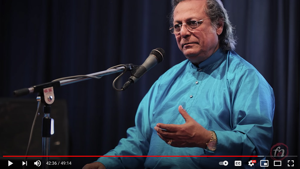 Swapan Chaudhuri sits behind a microphone in a turquiose garment, gesturing with one hand.