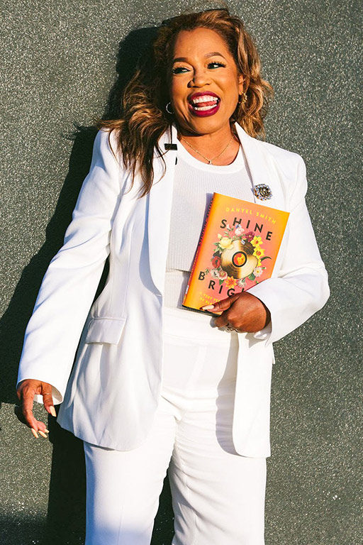 A color photograph of Danyel Smith, looking to their right, smiling a broad smile, dressed in a white suit, holding a copy of their book with a peach colored cover with flowers, gold record and text, standing in front of a concrete wall. 