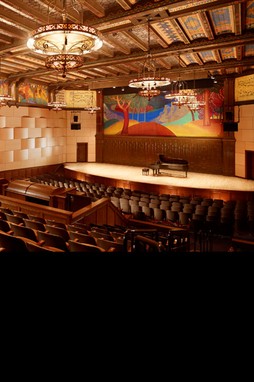 A color photograph of the Littlefield Concert Hall, with colorful frescoes around the ceiling and ornate chandeliers hanging down. A grand piano sits on stage.