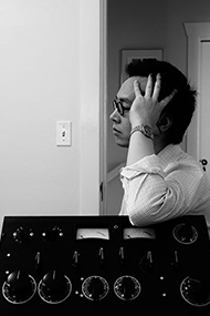 James Fei stands behind a modular synthesizer, with their body resting against the machine. Their hand supports their head, as the look in profile out of frame.