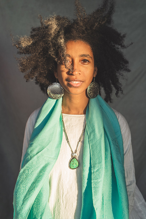 Alexis Pauline Gumbs looks into the camera, dressed in a white top and turquoise scarf, and large silver circular earrings, a golden light shines on their face, smiling. Photo courtesy of the author.