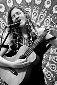 Maishi Lani sings into a microphone with their eyes closed while playing guitar, standing in front of a radial tapestry hanging. Phot Courtey of the Artist