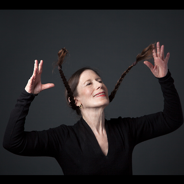 Meredith Monk flips her braids up into the air with a gentle smile on her face, eyes closed. She is dressed in a black top and in front of a grey background. Photo by Christine Alicino