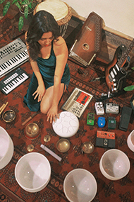 Composer maryzelle Ungo sits in their knees surrounded by a radial array of instruments. Photo courtesy of the Artist.