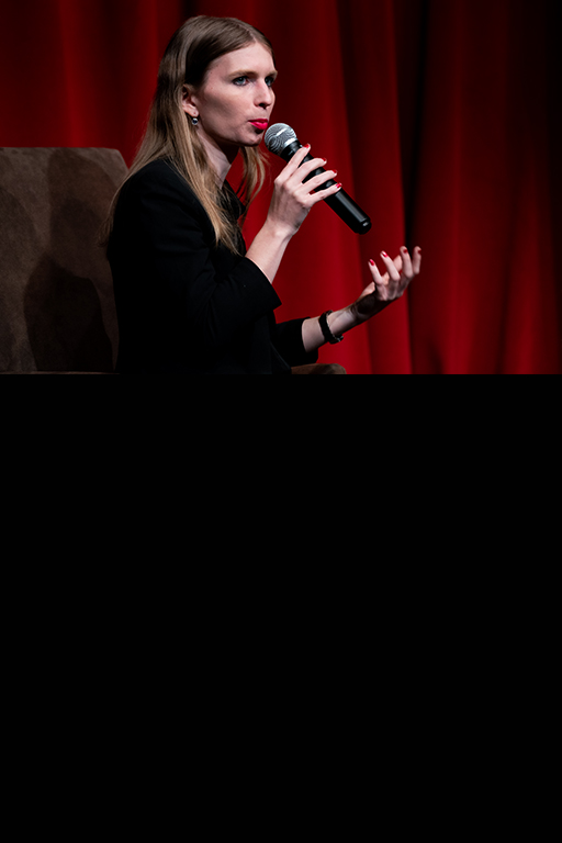 Chelsea Manning, speaking into a microphone to their mouth, dressed in a black shirt, their long shoulder length dark bonde hair resting on their shoulders, seated in front of red velvet curtain. Photo by Robbie Sweeny.