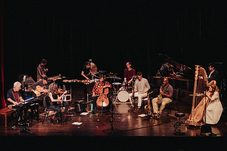 An improvisational ensemble of including keyboards, harp, violin, cello, drum set, saxophone are arrayed across a stage for a Mills Music Now concert in Oakland, California. Photo by Robbie Sweeny