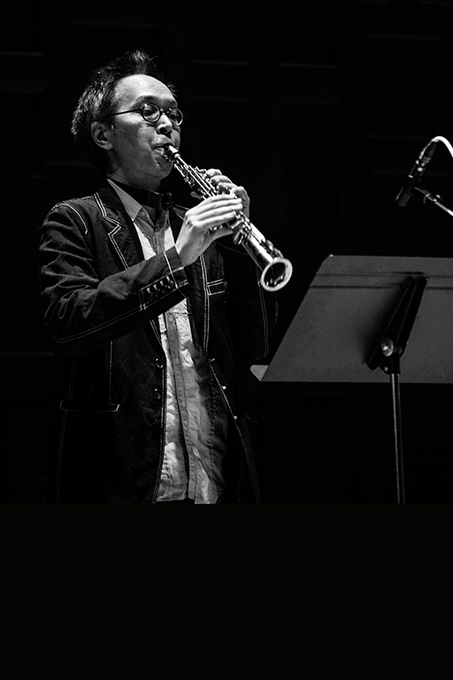 A black and white photograph of James Fei playing soprano saxophone, standing at a microphone with a music stand before them, head positioned slightly upward and eyes cast down through glasses, dressed in a black jacket and white shirt