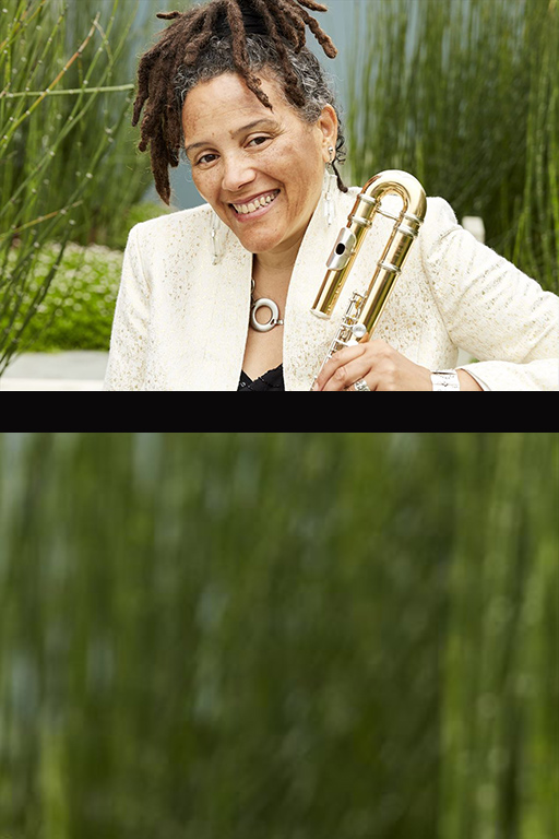 Composer and Improviser Nicole Mitchell smiles broadly into the camera, dressed in a cream and gold flecked blazer while holder a flute and seated before a garden of green equisetum. Photo by Amanda E Friedman.