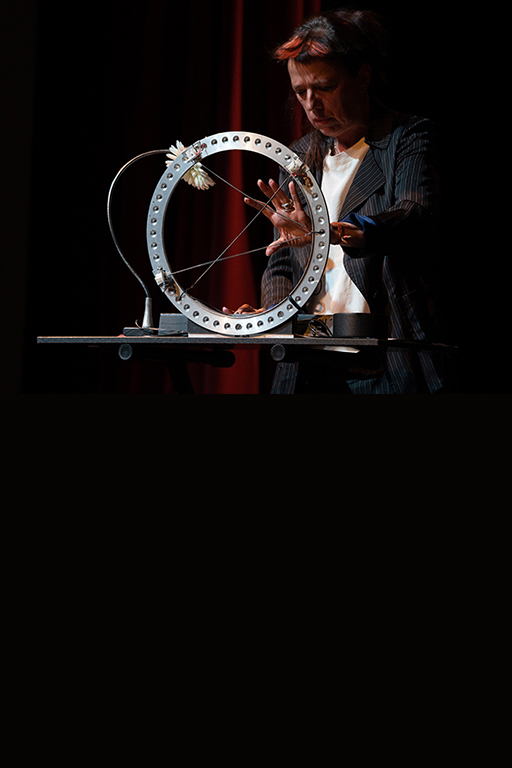 A color photograph of Laetitia Sonami, dressed in a dark suit coat and dark shirt, playing a built electronic musical instrument consisting of a standing metal circle with three wires crossing the center opening. Photo by Robbie Sweeny.