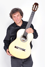 Guitarist David Tanenbaym is photographed in a studio, dressed in a black shirt in a white field. The guitar is held in front of the chest. Photo courtesy of the artist. 
