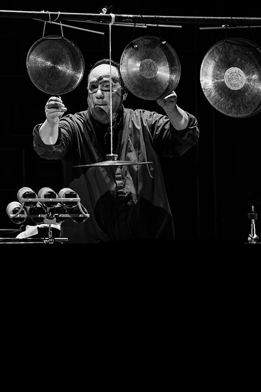 A black and white photo of William Winant playing percussion surrounded by an array of instruments including cymbals and gongs handing from a structure. Photo by Robbie Sweeny.