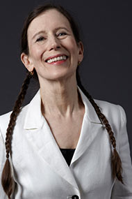 Meredith Monk smiles to the camera. Her hair in distinctive long braids, she is dressed in a white blazer.