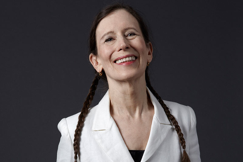 eredith Monk smiles wearing a white coat and braids, standing before a dark grey backdrop.