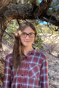 Tova Strong is photographed outside standing under a tree branch, dressed in a red and white plaid shirt. Their long brown hair over one shoulder, wearing dark rimmed glasses and a gentle smile. Photo Courtesy of Tovah Strong.