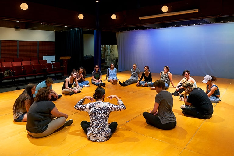 A group of performing artists are seated in a circle on the stage of Mills College's Rothwell Theater, which is available for community rentals.